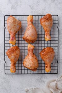 Six seasoned raw chicken drumsticks on a drying rack.