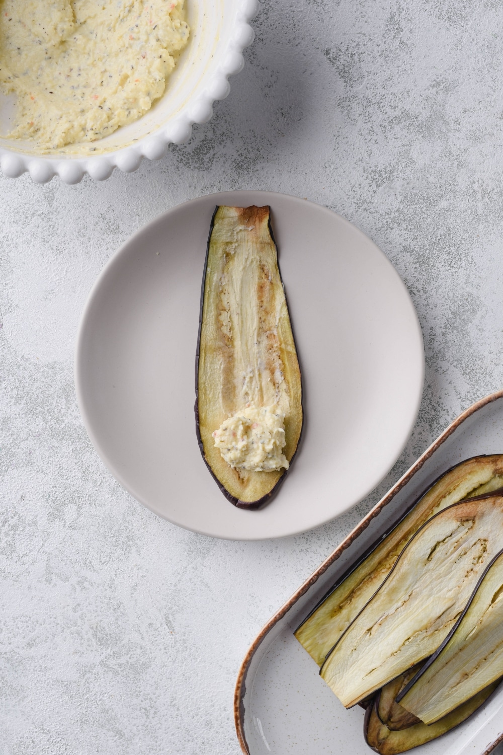 Thinly sliced eggplant on a plate with a spoonful of filling, next to a bowl of filling and a plate of eggplant slices.