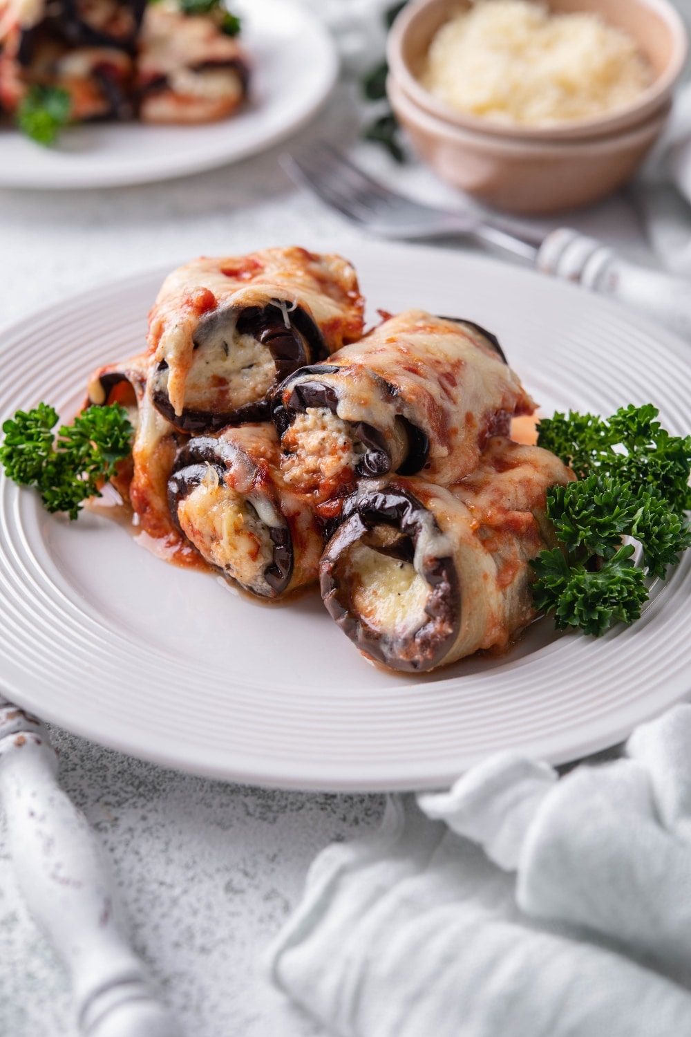 Eggplant rollatini served on a plate with curly parsley, in front of a second plate of eggplant rollatini and a bowl of shredded cheese.