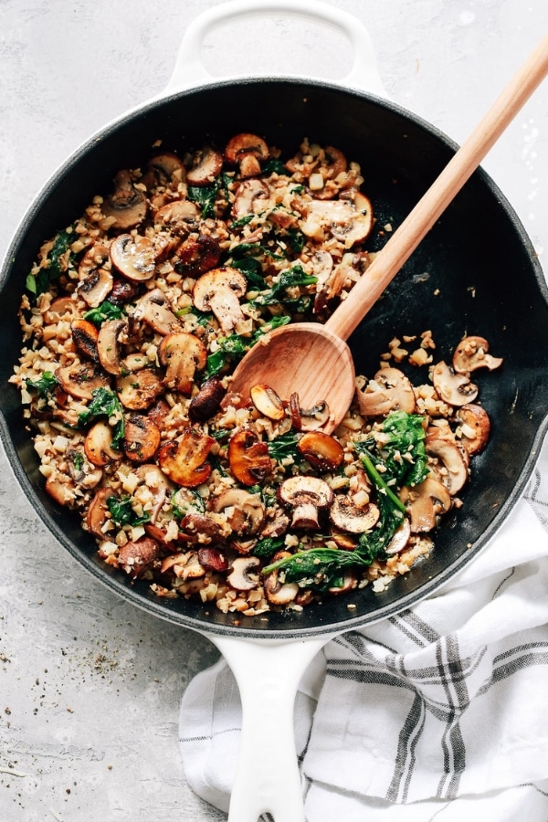 A wooden spoon in a skillet that is filled with mushrooms and cauliflower rice.