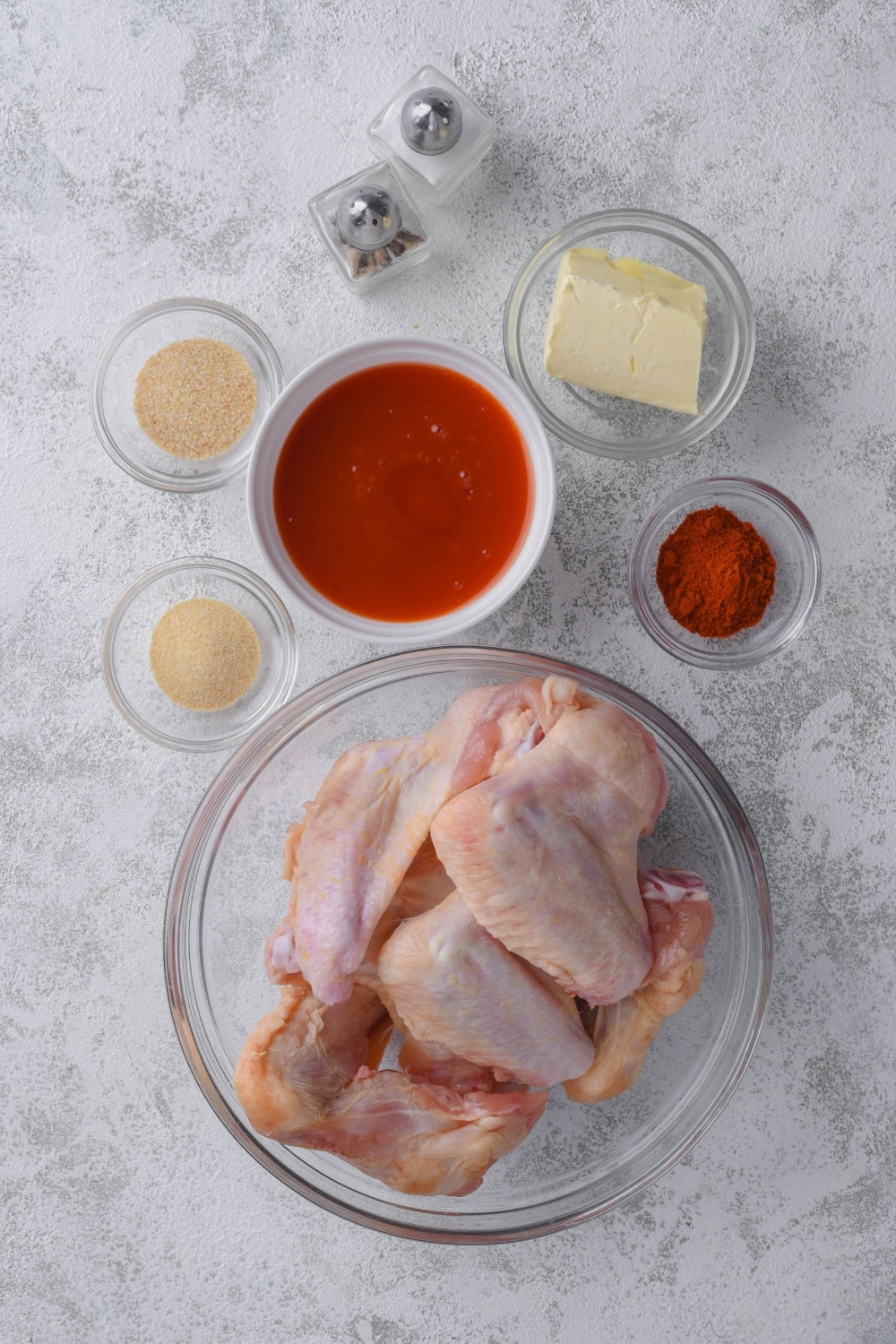 Raw chicken wings in a glass bowl, a bowl of hot sauce, small bowls of butter, paprika, garlic powder, and baking powder, and salt and pepper shakers.