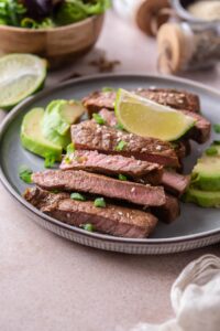 Sirloin tip steak on a plate, sliced and served with avocado slices and lime wedges, garnished with chopped green onions and sesame seeds.