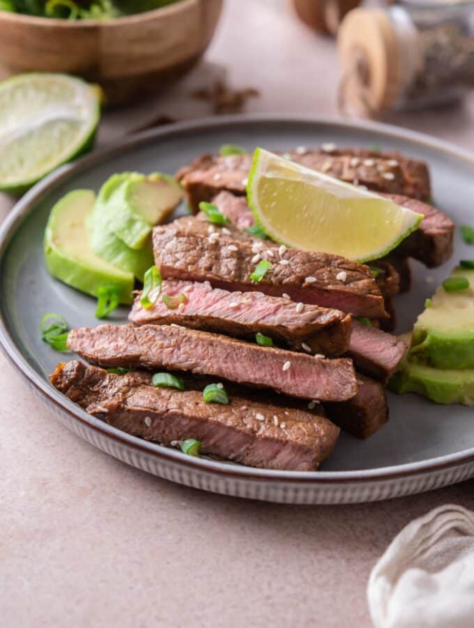 Sirloin tip steak on a plate, sliced and served with avocado slices and lime wedges, garnished with chopped green onions and sesame seeds.