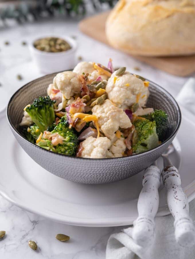 Broccoli and cauliflower salad mixed with dressing and topped with pumpkin seeds and cheddar cheese in a grey bowl set over a white plate. In the back is a loaf of bread and a small bowl of seeds.