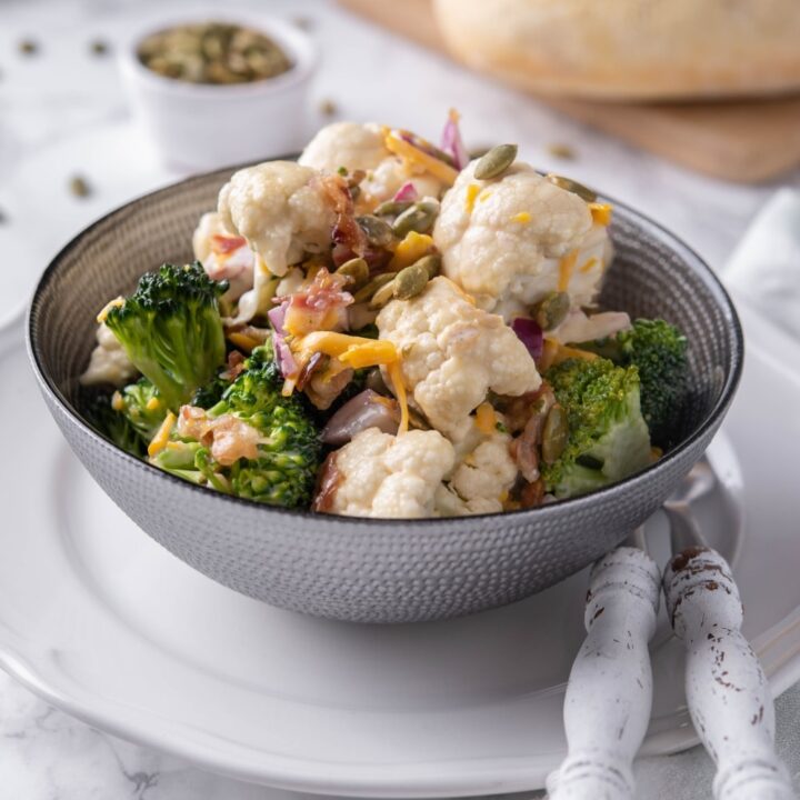 Broccoli and cauliflower salad mixed with dressing and topped with pumpkin seeds and cheddar cheese in a grey bowl set over a white plate. In the back is a loaf of bread and a small bowl of seeds.