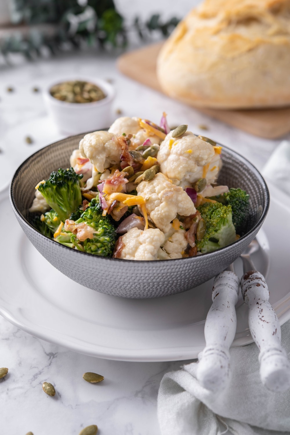 Broccoli and cauliflower salad mixed with dressing and topped with pumpkin seeds and cheddar cheese in a grey bowl set over a white plate. In the back is a loaf of bread and a small bowl of seeds.