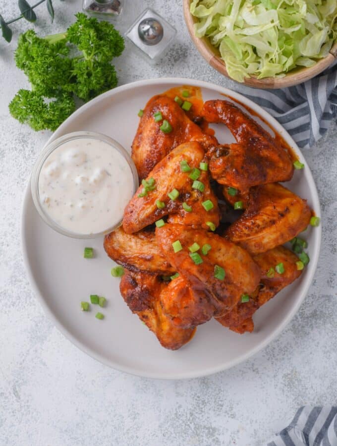 Top view of baked chicken wings topped with chopped green onions and served with a small bowl of ranch. Part of a bowl of shredded lettuce is on the side.