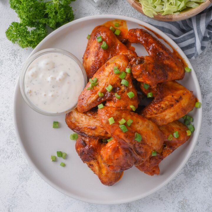 Top view of baked chicken wings topped with chopped green onions and served with a small bowl of ranch. Part of a bowl of shredded lettuce is on the side.