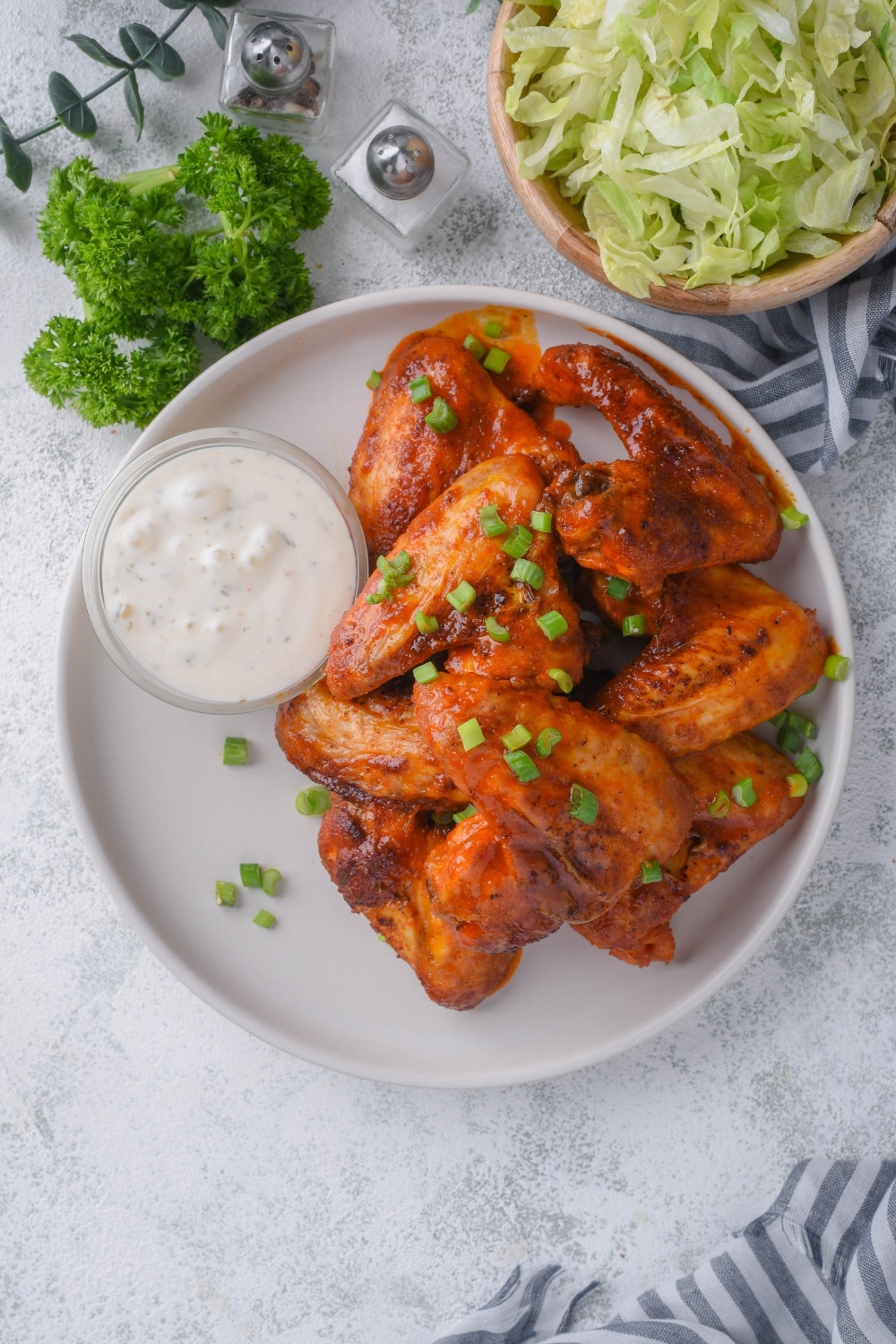 Top view of baked chicken wings topped with chopped green onions and served with a small bowl of ranch. Part of a bowl of shredded lettuce is on the side.