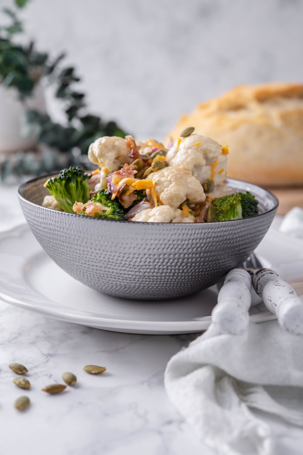 Broccoli and cauliflower salad mixed with dressing and topped with pumpkin seeds and cheddar cheese in a grey bowl set over a white plate. In the back is a loaf of bread.