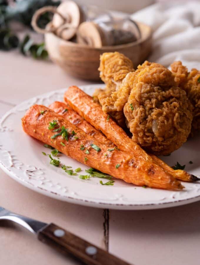 Grilled carrots garnished with parsley and served with fried chicken on a white plate.