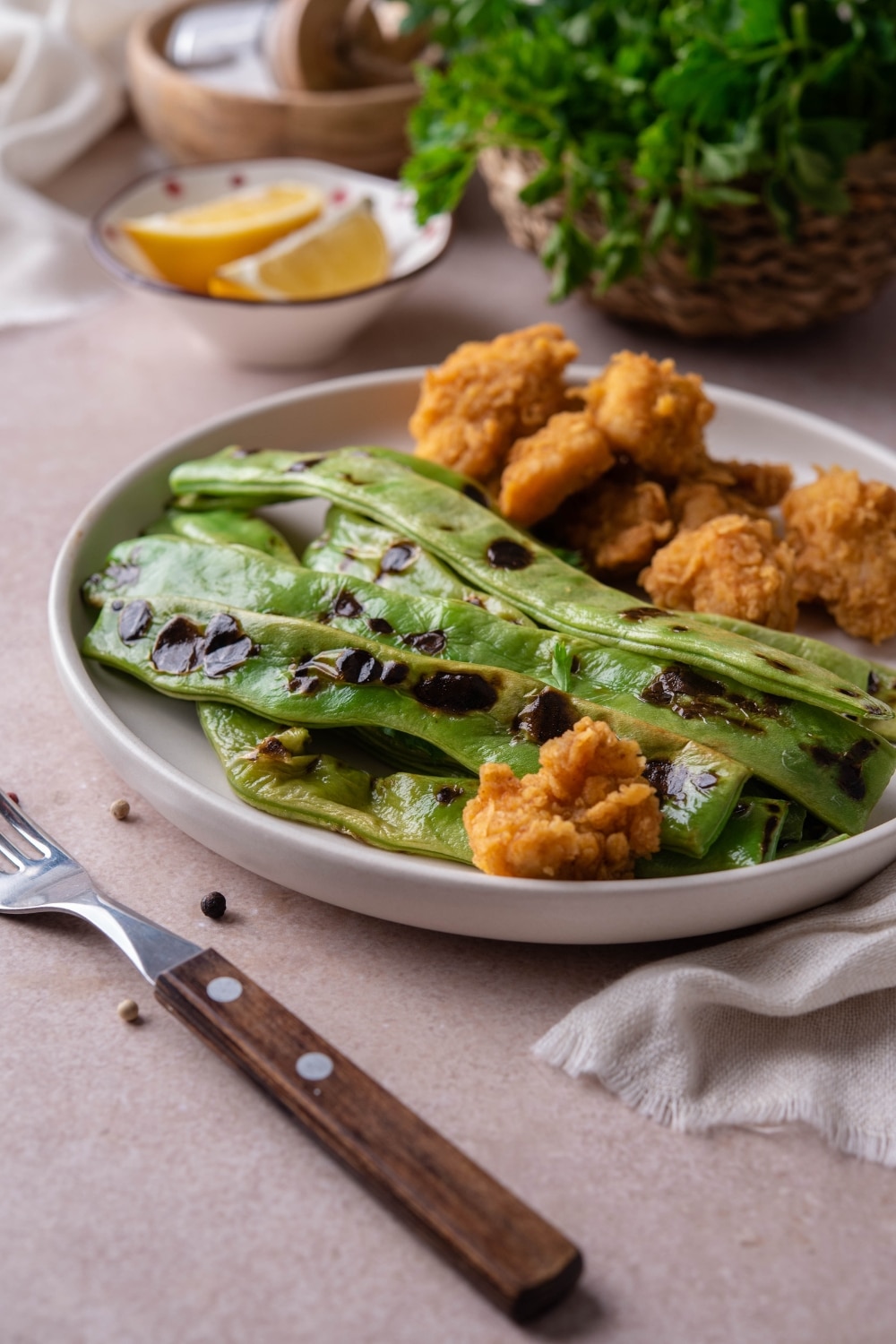 Grilled green beans served with chicken nuggets on a white plate, with a small bowl of lemon wedges on the side.