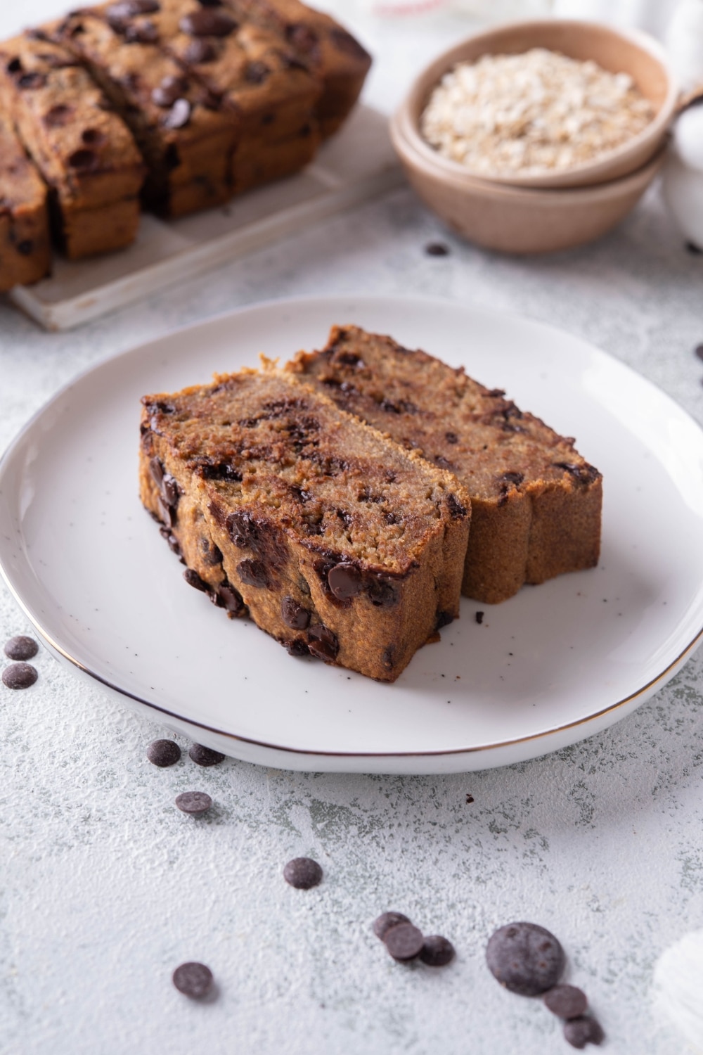 Two slices of chocolate chip oatmeal banana bread on a plate. Behind it is a plate of the rest of the banana bread and a small bowl of oats.