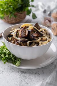 Roasted eggplant mixed with fettucine pasta in a white bowl, served over a plate garnished with fresh parsley.