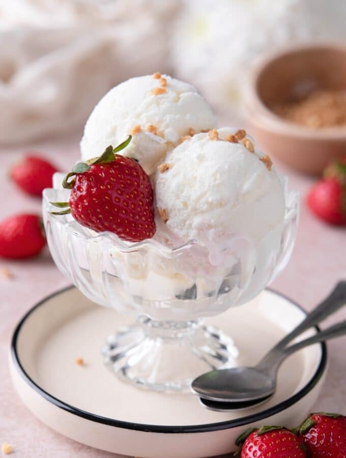 Two scoops of frozen yogurt ice cream with a strawberry between them in a glass bowl on a white plate.