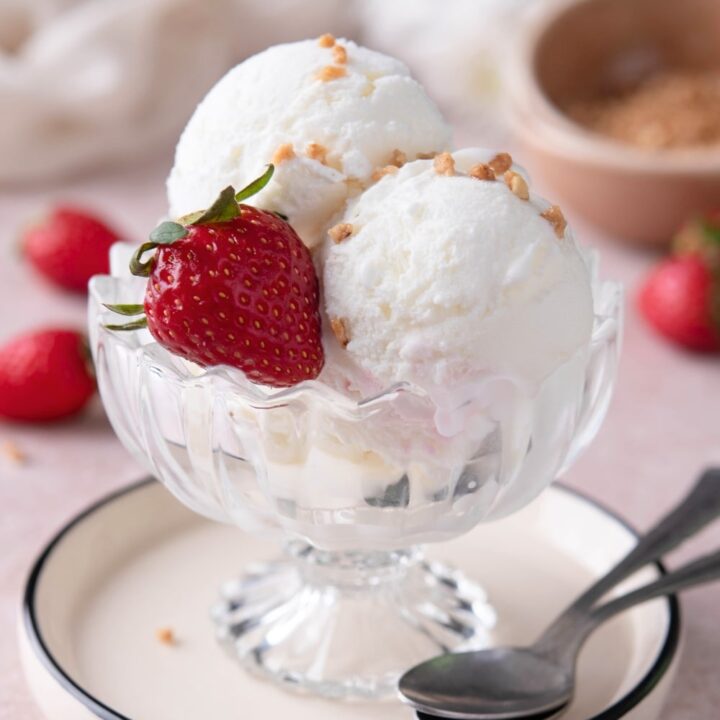 Two scoops of frozen yogurt ice cream with a strawberry between them in a glass bowl on a white plate.