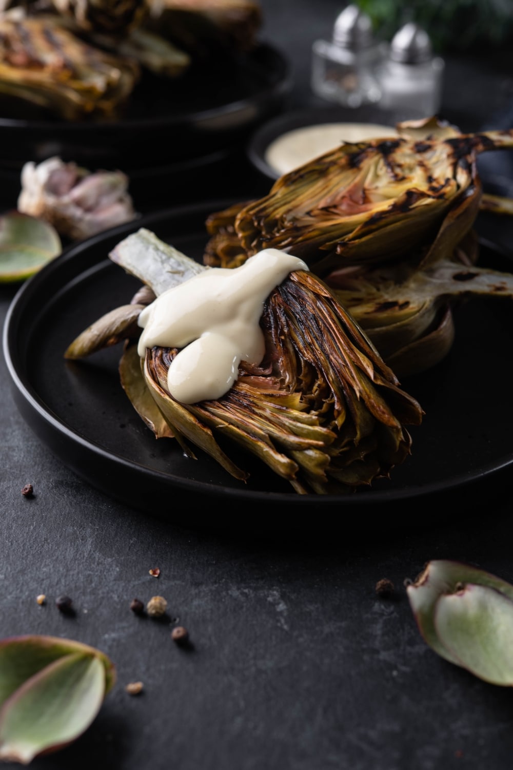 Grilled artichokes drizzled with aioli on a black plate.