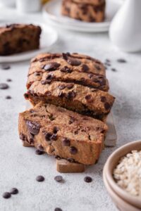 A sliced loaf of chocolate chip oatmeal banana bread on a wood cutting board. Behind are more plate of banana bread slices.