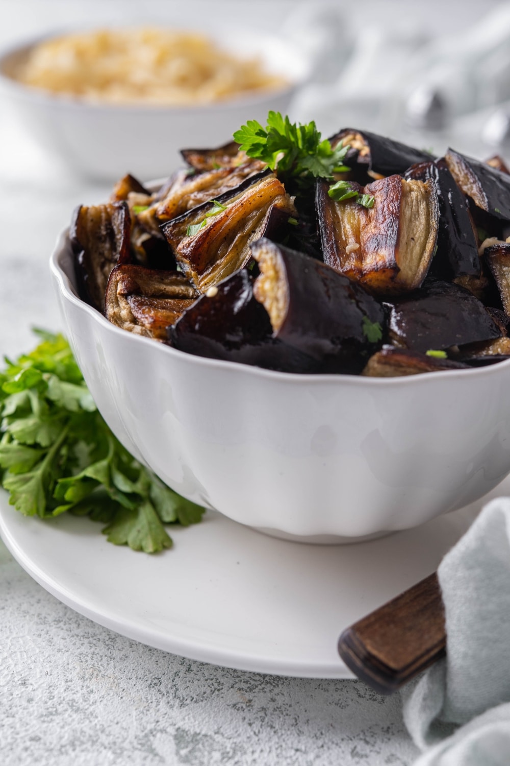 Roasted eggplant garnished with fresh parsley in a white bowl set over a plate.