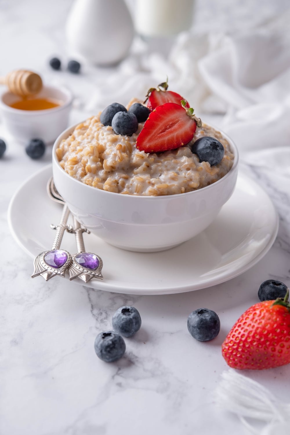 Steel cut oatmeal topped with blueberries and sliced strawberries, served in a white bowl set over a plate. Behind is a small bowl of honey with a honey dipper.