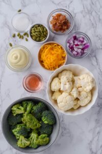A bowl of broccoli florets, a bowl of cauliflower florets, and small bowls of pumpkin seeds, chopped red onions, bacon bits, mayonnaise, red wine vinegar, and sugar.