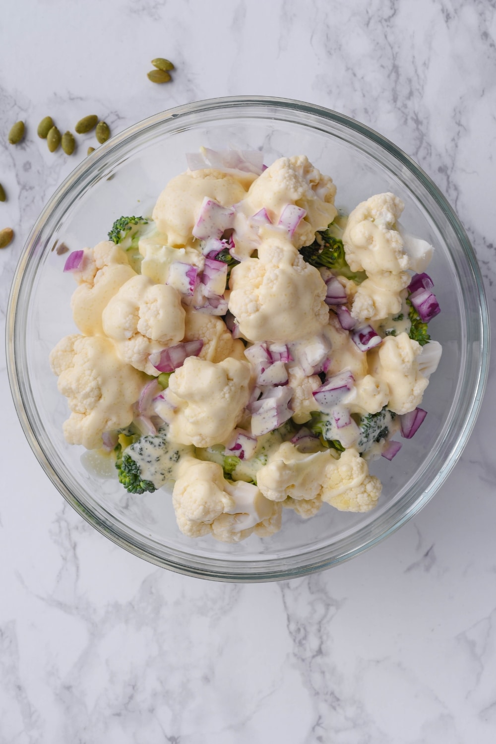 Cauliflower and broccoli florets with chopped red onions, coated in creamy salad dressing, in a glass bowl.