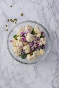 Cauliflower and broccoli florets with chopped red onions in a glass bowl.
