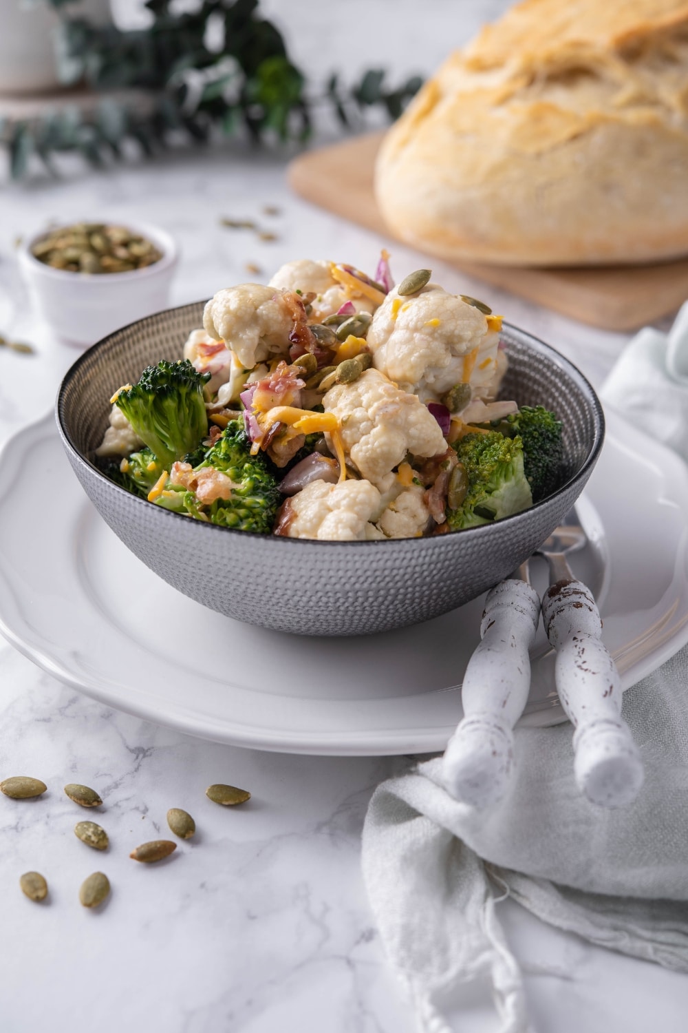 Broccoli and cauliflower salad topped with pumpkin seeds and cheddar cheese served in a grey bowl. Behind is a loaf of bread and a small bowl of seeds.