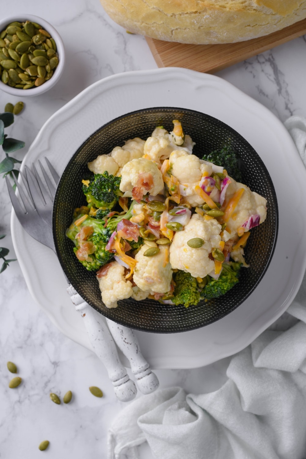 Top view of broccoli and cauliflower salad. It's topped with pumpkin seeds, bacon bits, and cheddar cheese and served with a loaf of bread and a small bowl of seeds.