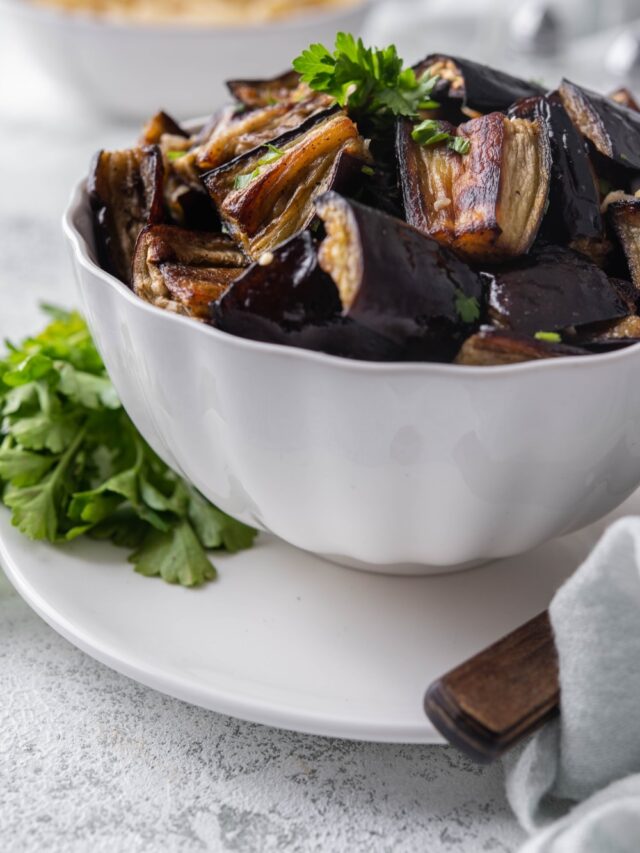 Roasted eggplant garnished with fresh parsley in a white bowl set over a plate.