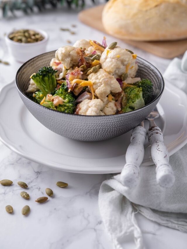 Broccoli and cauliflower salad topped with pumpkin seeds and cheddar cheese served in a grey bowl. Behind is a loaf of bread and a small bowl of seeds.