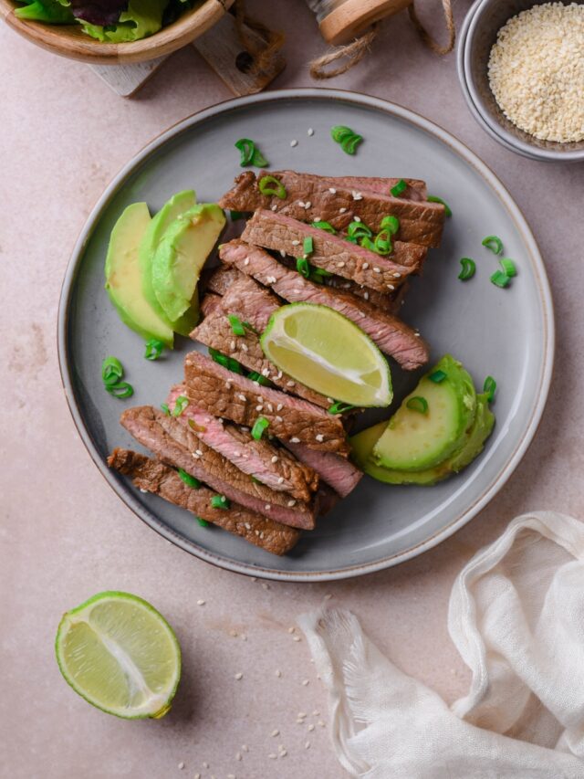 Sliced medium sirloin tip steak on a plate served with avocado slices and garnished with lime wedges and chopped green onion.