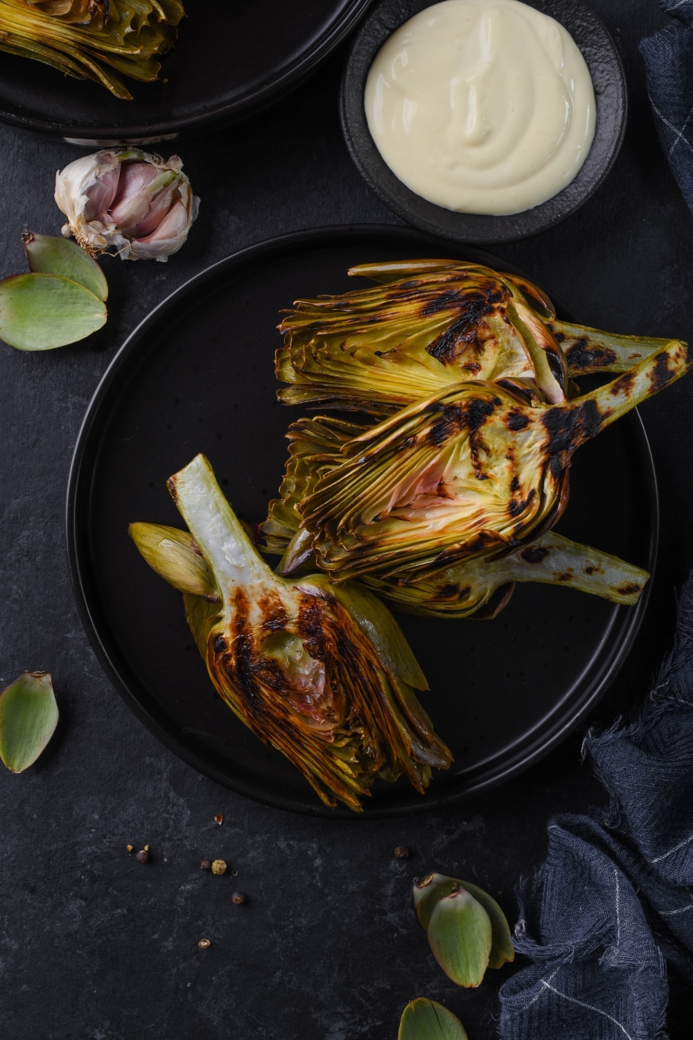 Top view of grilled artichokes on a black plate served with a saucer of aioli.