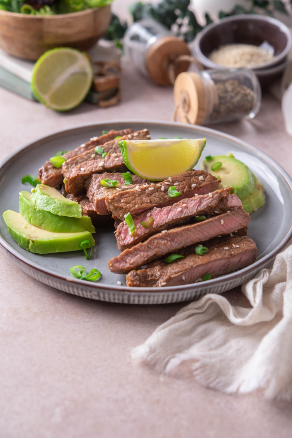 Sliced sirloin tip steak on a plate garnished with avocado slices, lime wedges, chopped green onions, and sesame seeds.