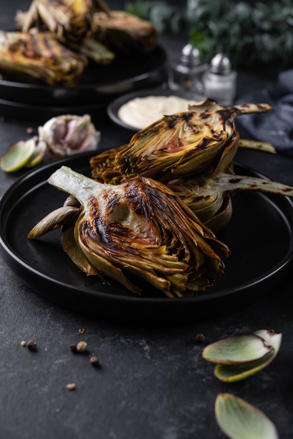 A plate of grilled artichokes next to a saucer of aioli, fresh garlic bulbs, and another plate of grilled artichokes.