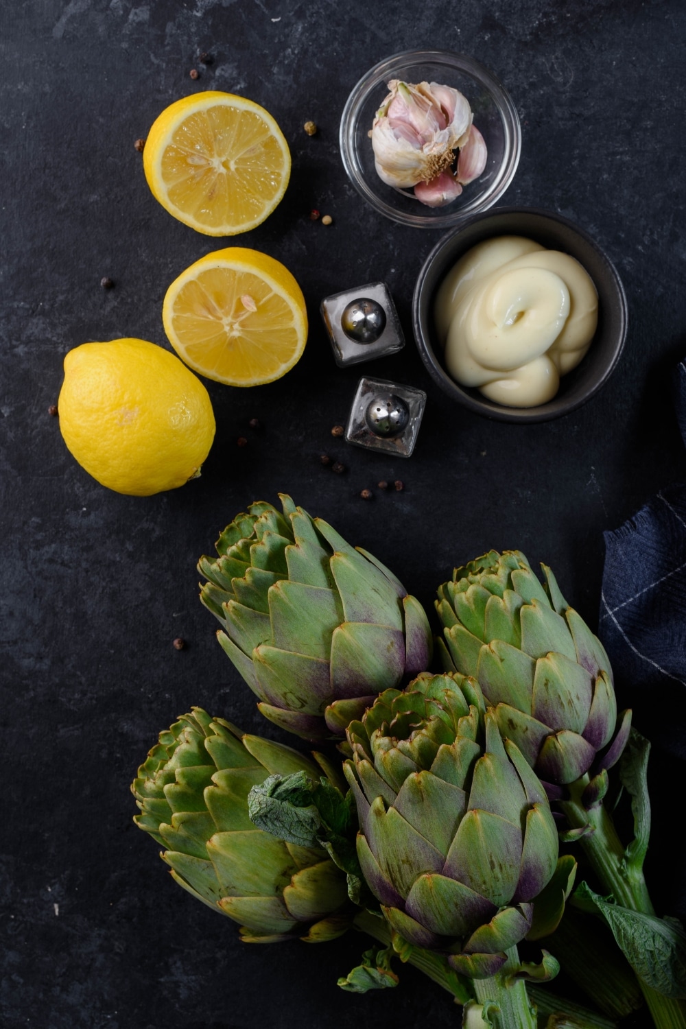 Four fresh artichokes, lemons, a small bowl of mayonnaise, a small bowl of garlic bulbs, and salt and pepper shakers.