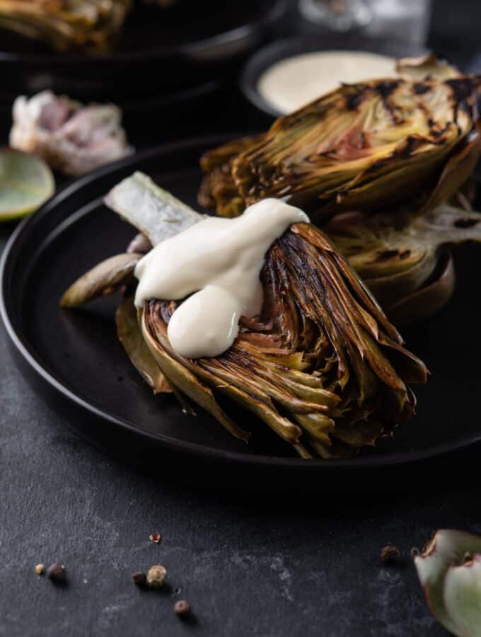 Three grilled artichokes on a black plate, one drizzled with aioli.