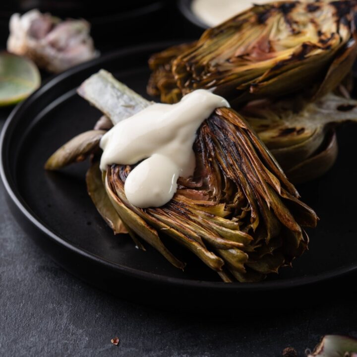 Three grilled artichokes on a black plate, one drizzled with aioli.