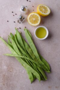 Raw green beans, a small bowl of oil, a halved lemon, and salt and pepper shakers.