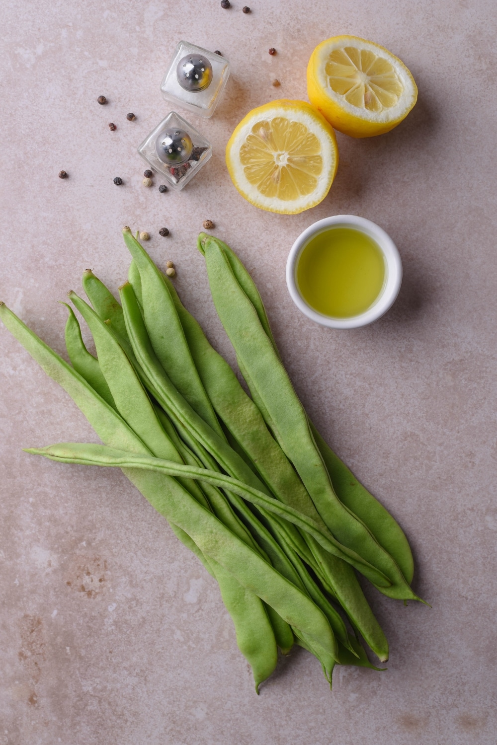 Raw green beans, a small bowl of oil, a halved lemon, and salt and pepper shakers.