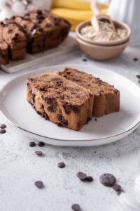 A plate with two slices of chocolate chip oatmeal banana bread. Behind is the rest of the banana bread and a small bowl of oats.