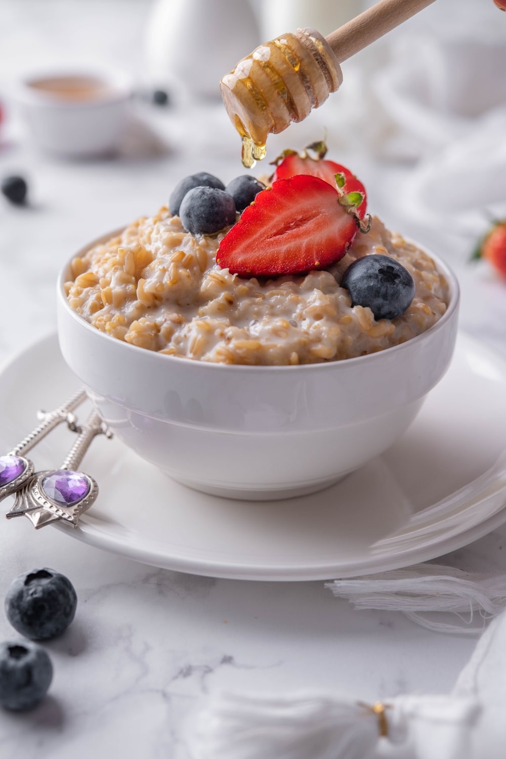 A honey dipper dripping honey onto a bowl of creamy steel cut oats garnished with sliced strawberries and blueberries.