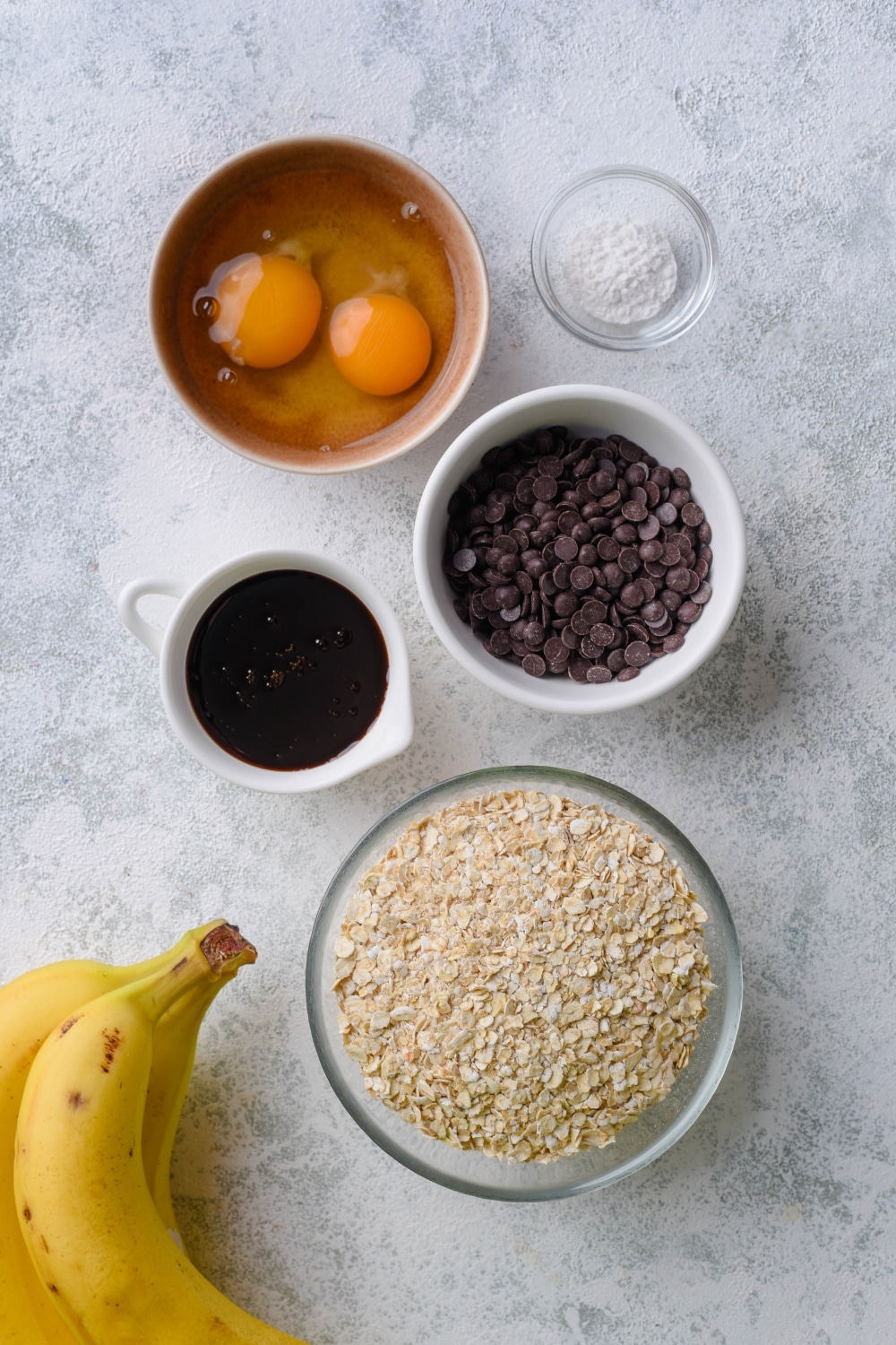 Fresh bananas, bowls of dry oats, egg, chocolate chips, baking soda, and a mini pitcher of date syrup.