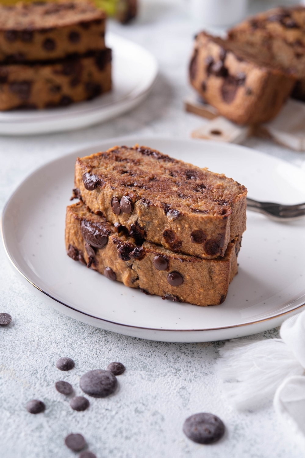 Two oatmeal chocolate chip banana bread slices stacked on a white plate with a fork. In the back are more banana bread slices.