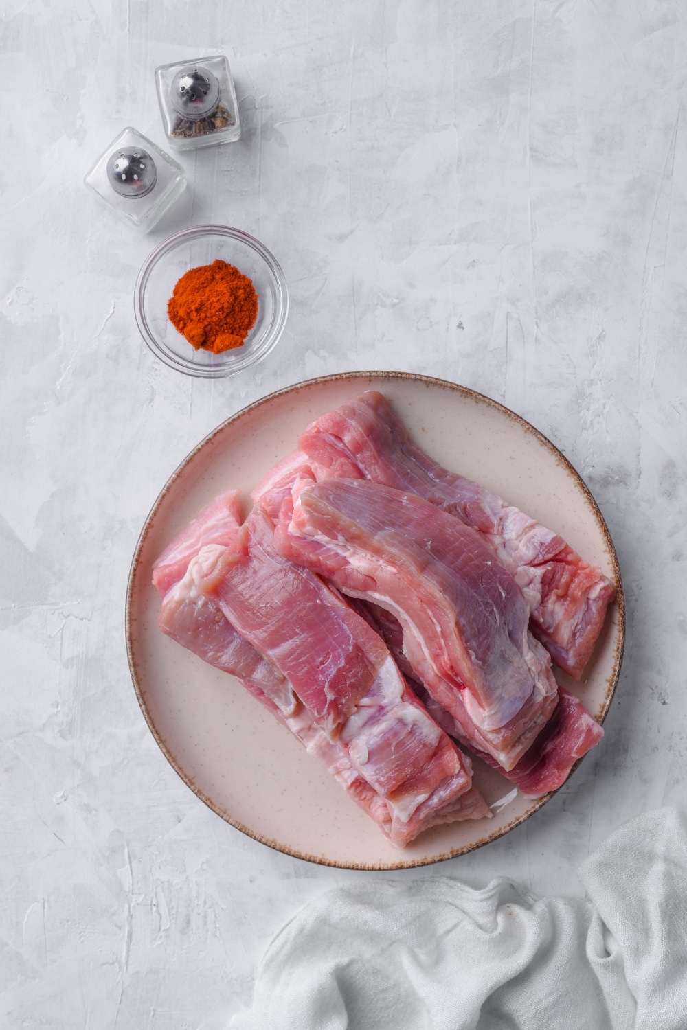 Thick pork belly strips on a plate, a small glass bowl of paprika, and salt and pepper shakers.