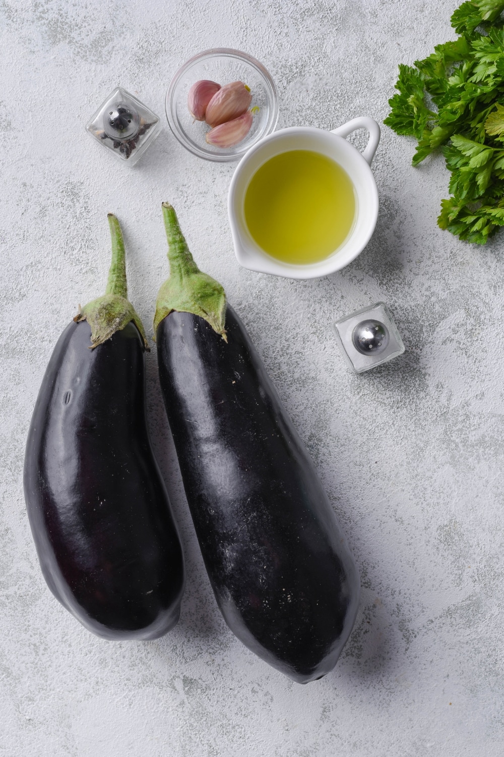 Two eggplants, a small pitcher of olive oil, a small bowl with three cloves of garlic, a bunch of parsley, and salt and pepper shakers.