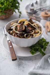 Roasted eggplant and fettucine mixed in a bowl, served over a plate with fresh parsley and a fork.