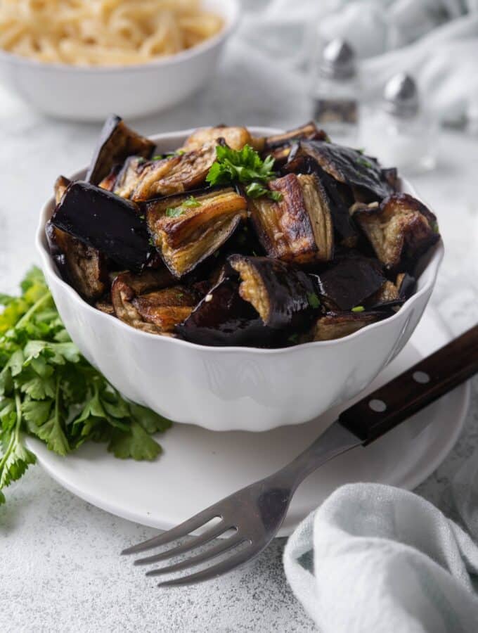 A bowl of roasted eggplants garnished with parsley, served over a plate with a fork. In the back is a bowl of fettucine pasta.