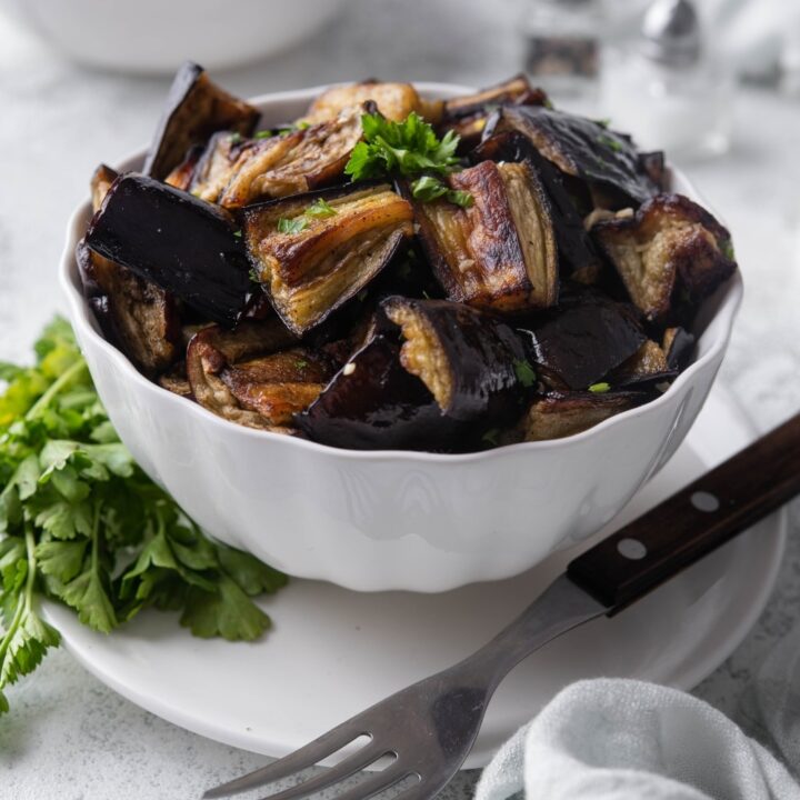 A bowl of roasted eggplants garnished with parsley, served over a plate with a fork. In the back is a bowl of fettucine pasta.