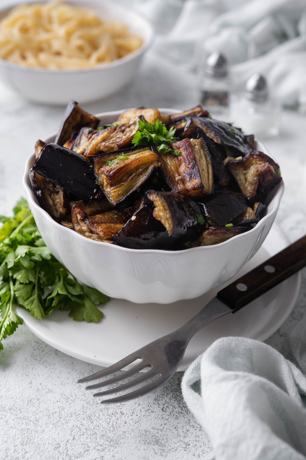 A bowl of roasted eggplants garnished with parsley, served over a plate with a fork. In the back is a bowl of fettucine pasta.